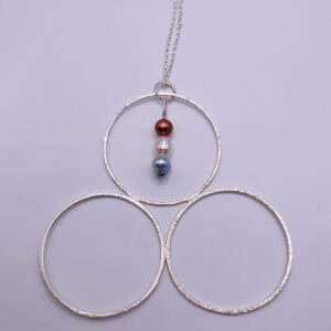 Unique handmade three large textured hoops joined to look like a three leaf clover. The pendant is made from recycled sterling silver. Three coloured cultured pearls hang down from the top hoop, one grey, one cream and one red. The beautiful pendant hangs from a silver trace chain. On a white background.