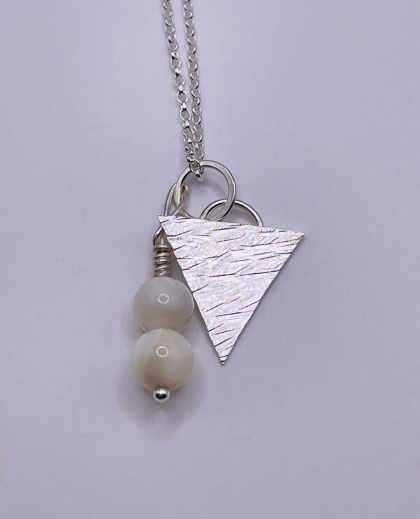 Attractive handmade pendant consisting of a small textured triangle made from recycled sterling silver as well as two cream mother of pearl beads which hang down next to the triangle. The pendant hangs on a silver trace chain. On a white background.